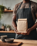 Kraft paper delivery bag with folded top held in hands; dark apron on person