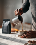 Metal kettle pouring dark coffee into a cup, hands holding a ceramic mug